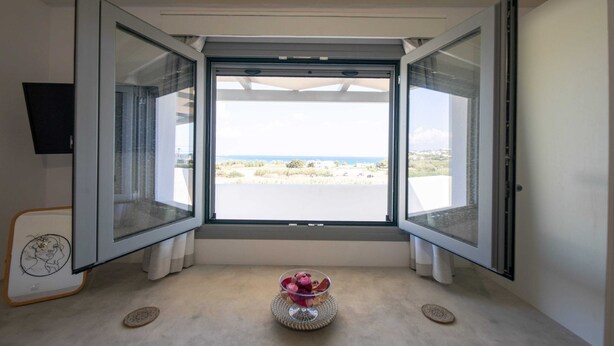 A room with a large sliding glass door leading to a beach view, a small table with a decorative bowl, and a framed picture on the wall. - Paros Beach Studio Casa Costa Oro, 300m from Golden Beach (1BR 1BA) (Paros)