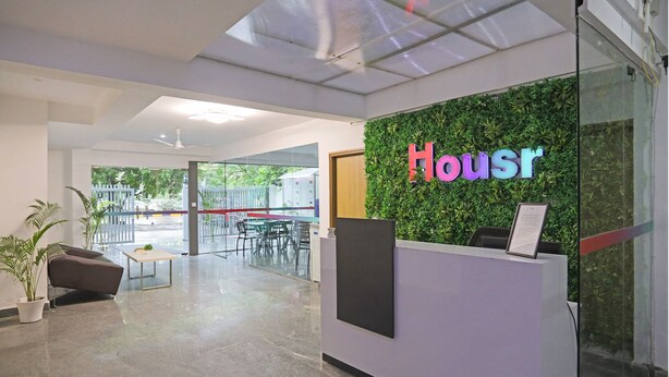 Área de recepção de hotel com parede verde e um letreiro 'Housr', balcão de atendimento, espaço de estar e porta de vidro que dá para o exterior.