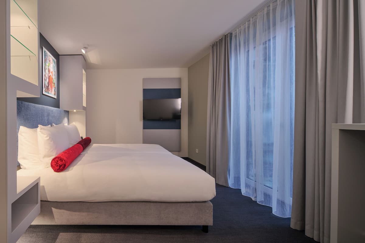 Une chambre d’hôtel moderne dotée d’un grand lit, d’un téléviseur à écran plat fixé au mur et d’une grande fenêtre avec des rideaux légers.