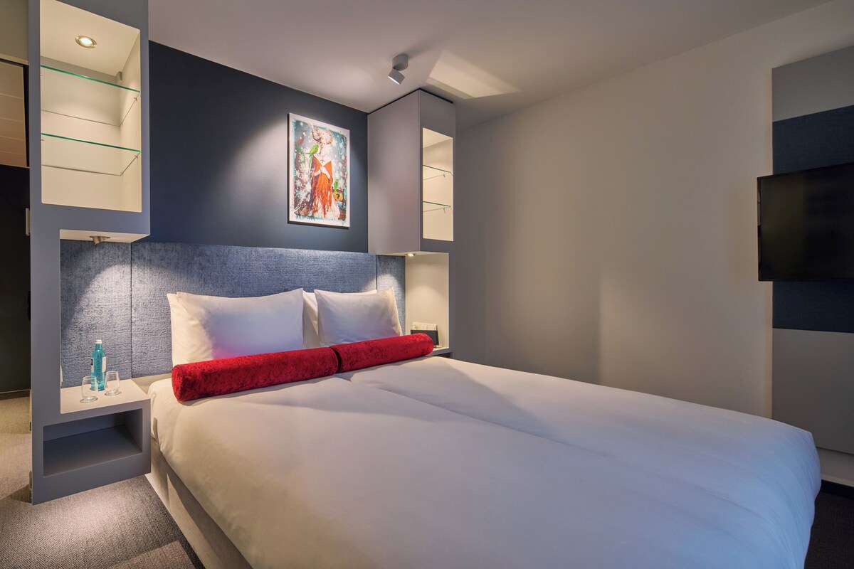 A modern hotel room with a large bed, a flat-screen TV, and a painting on the wall.