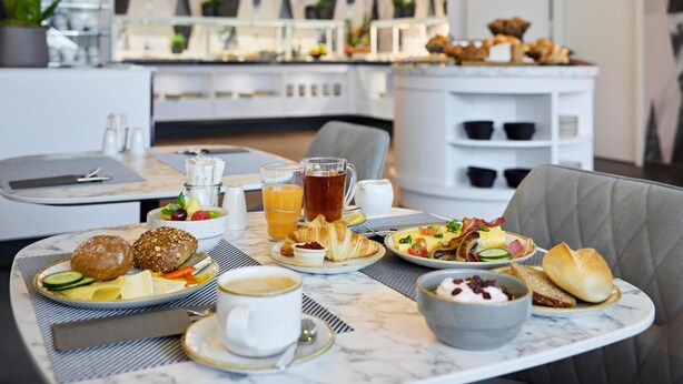 Una tavola imbandita con croissant, frutta e caffè, all'interno di una camera d'albergo moderna con vista sulla città.