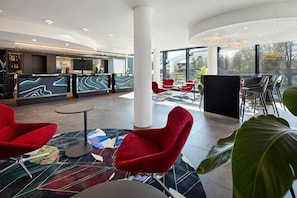 Un hall d’hôtel moderne avec un comptoir d’accueil, des fauteuils rouges, un coin bar et de grandes fenêtres donnant sur l’extérieur.