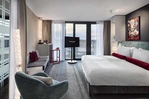Une chambre d’hôtel équipée d’un lit, d’un bureau, d’une chaise, d’une télévision et d’une grande fenêtre donnant sur la ville.