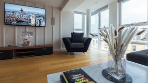 Ein modernes Wohnzimmer mit einem großen Flachbildfernseher, der eine Reisegesellschaft zeigt, einem gemütlichen Sofa, einem Couchtisch mit einem Buch und einer Vase mit Pampasgras.