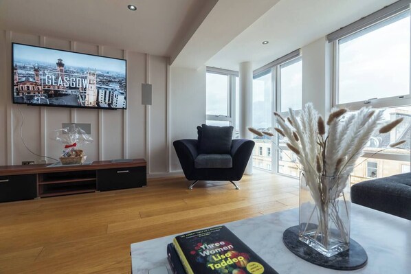 A modern living room with a large flat-screen TV displaying a travel show, a comfortable sofa, a coffee table with a book, and a vase with pampas grass.