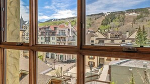 Vista a través de una ventana de un edificio de varios pisos, mostrando una combinación de edificios residenciales y comerciales, un cielo despejado y un paisaje montañoso al fondo.