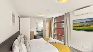 A hotel room with a bed, a bedside lamp, a window with curtains, a bathroom visible through an open door, and a painting on the wall.