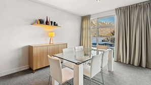 A modern hotel room with a glass dining table, white chairs, a wooden cabinet, a lamp, and a large window with a mountain view.