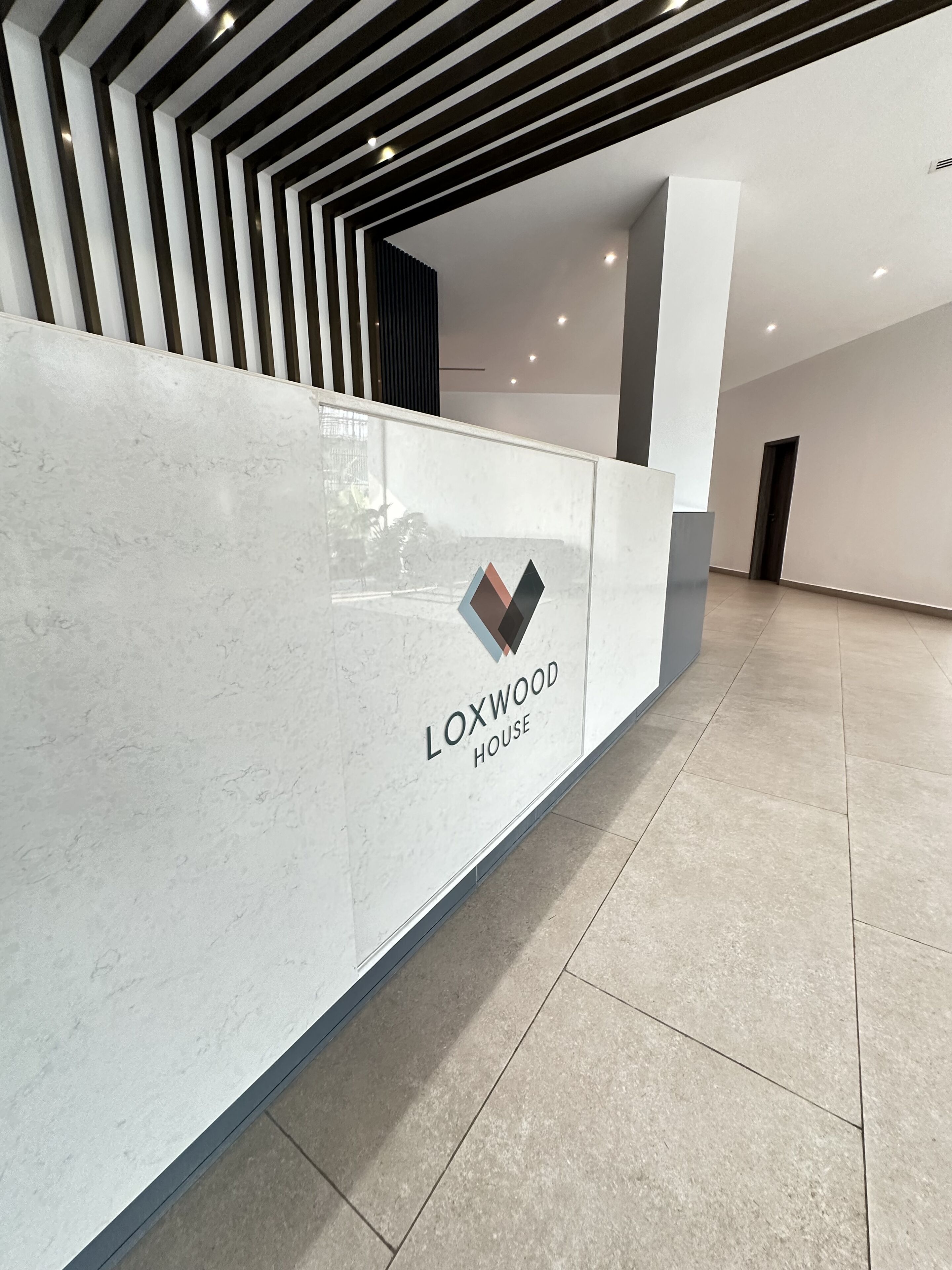 A modern reception desk with a logo and the name "LOXWOOD HOUSE" in a spacious lobby with a geometric ceiling design.