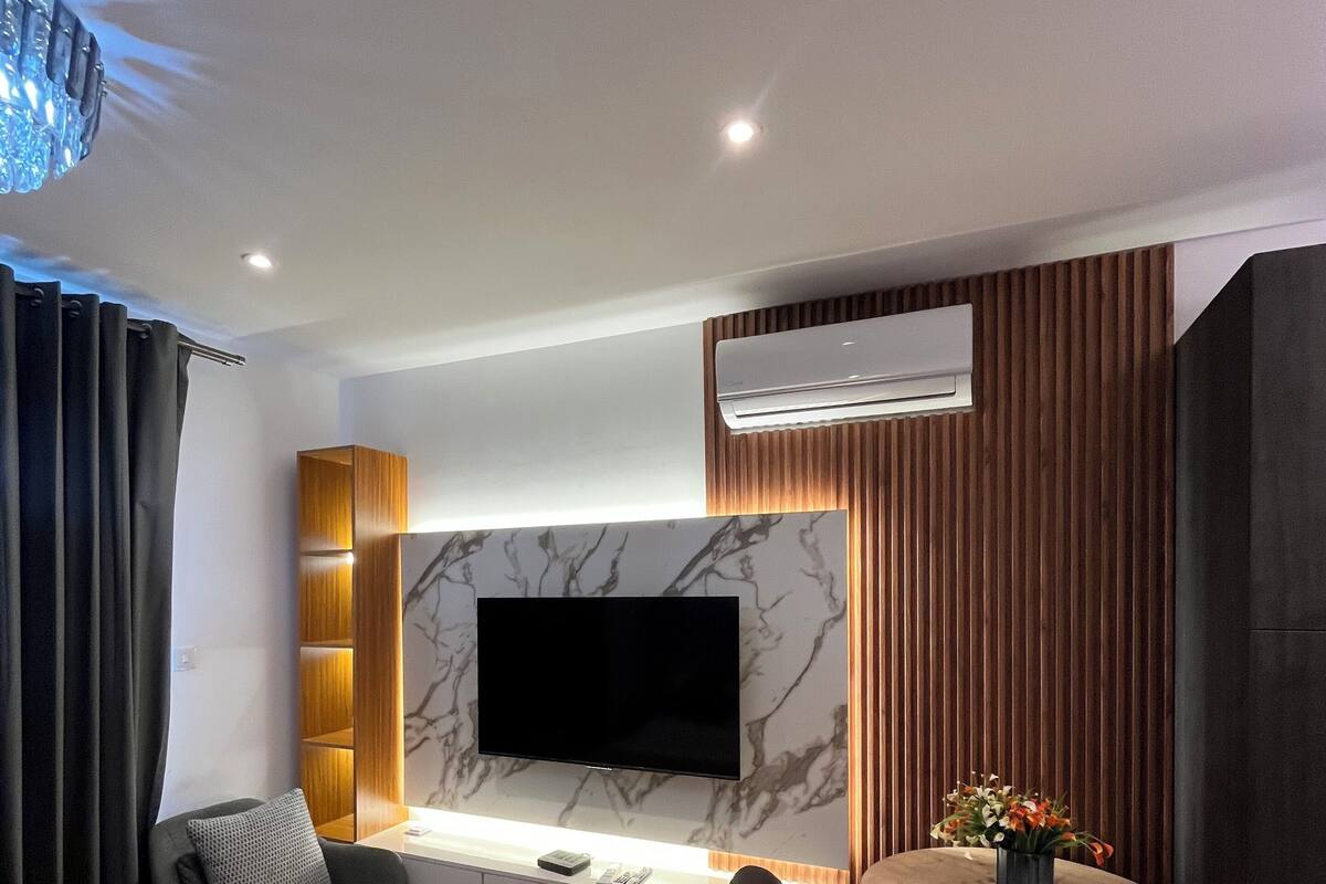 Quarto de hotel moderno com televisão de tela plana fixada em parede de mármore, detalhe em painel de madeira, mesa redonda de madeira com cadeiras, sofá com manta e unidade de ar condicionado.