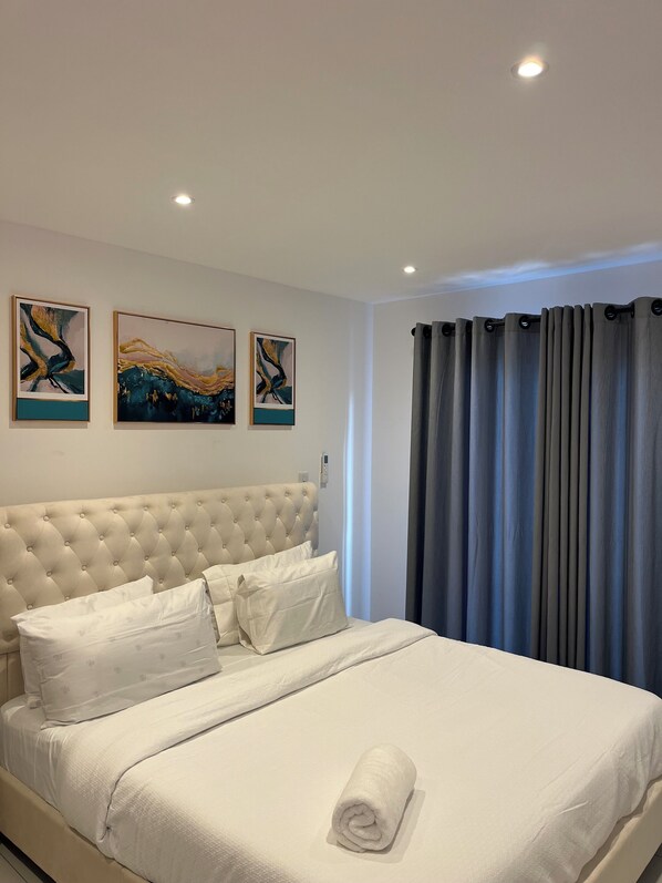 Un lit bien fait, avec une couette blanche et une serviette roulée, une tête de lit capitonnée et trois tableaux abstraits accrochés au mur.