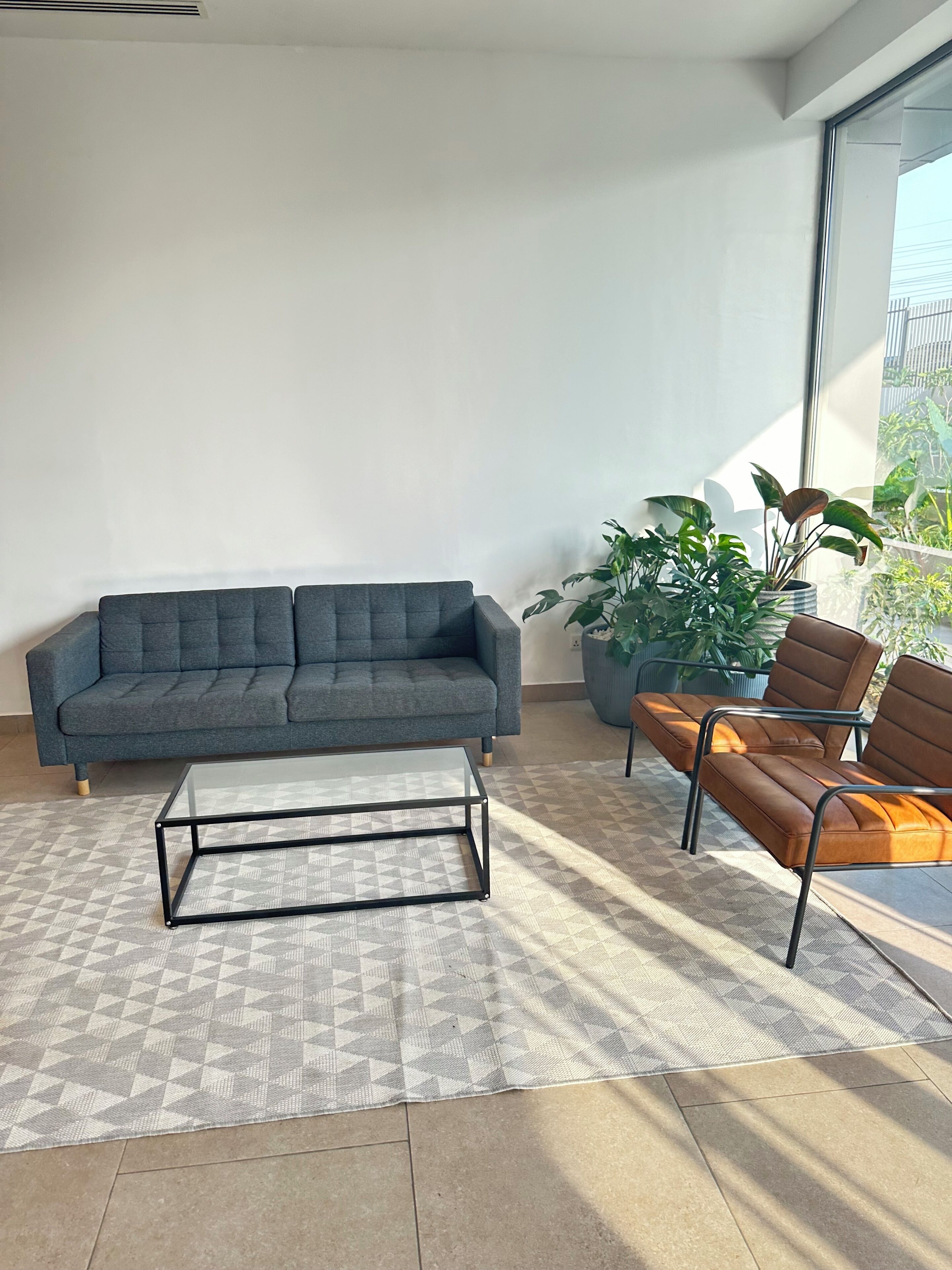 Ein modernes Wohnzimmer mit einem grauen Sofa, einem Glastisch und einem braunen Ledersofa. Übertöpfe und ein großes Fenster sorgen für natürliches Licht.