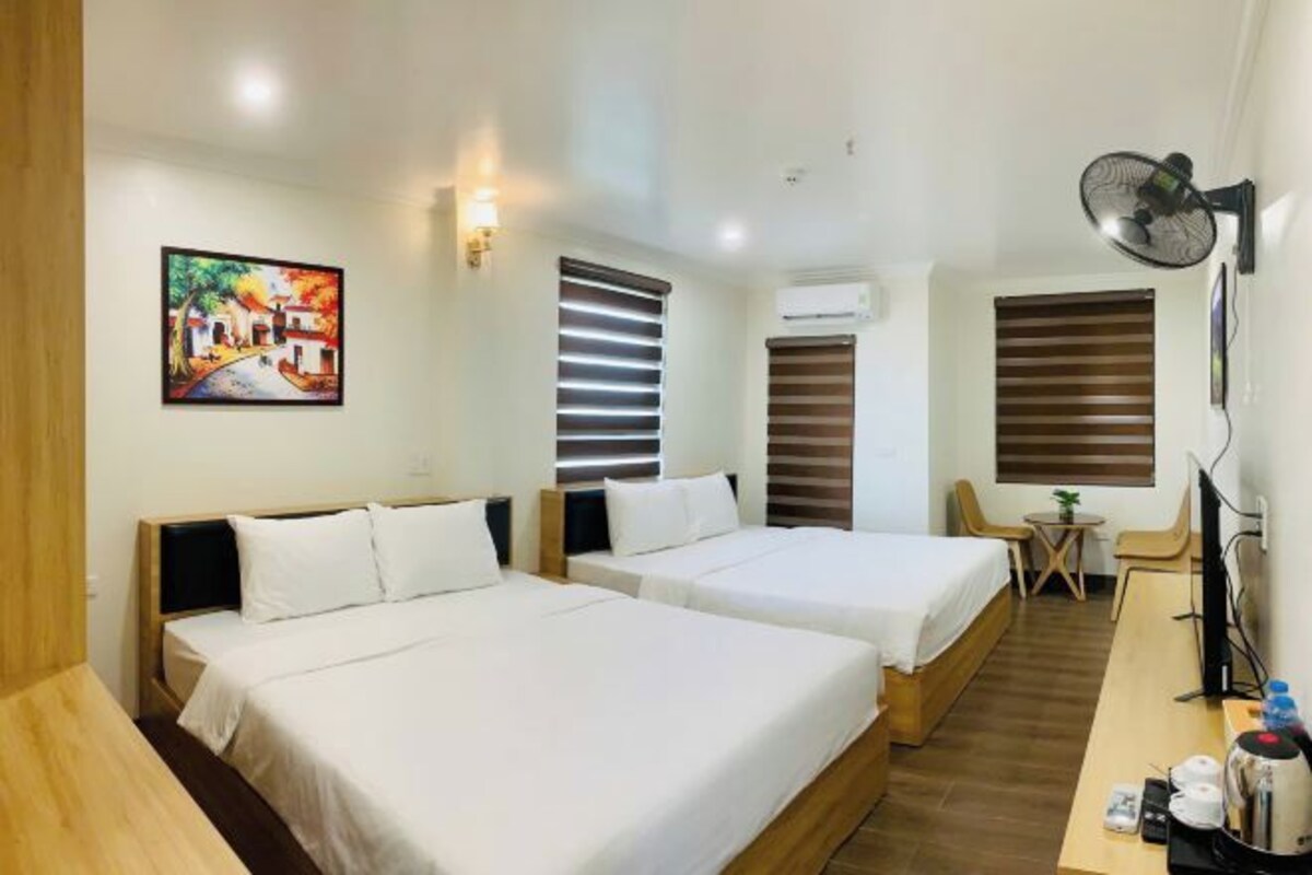 A hotel room with two beds, a desk, a chair, a small table, and a wall-mounted fan.