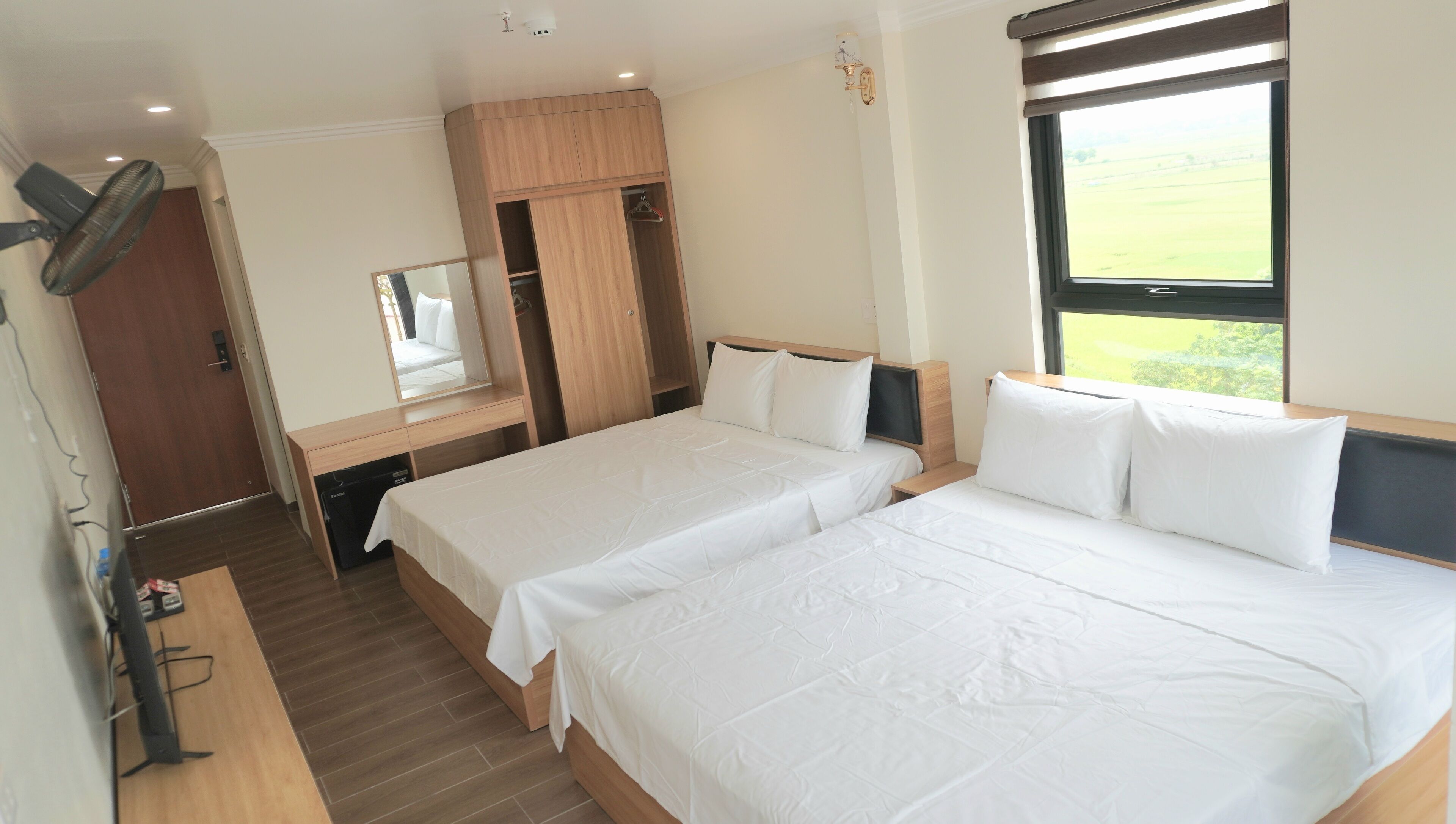 A hotel room with two beds, a wooden wardrobe, a mirror, and a window with a view of the countryside.