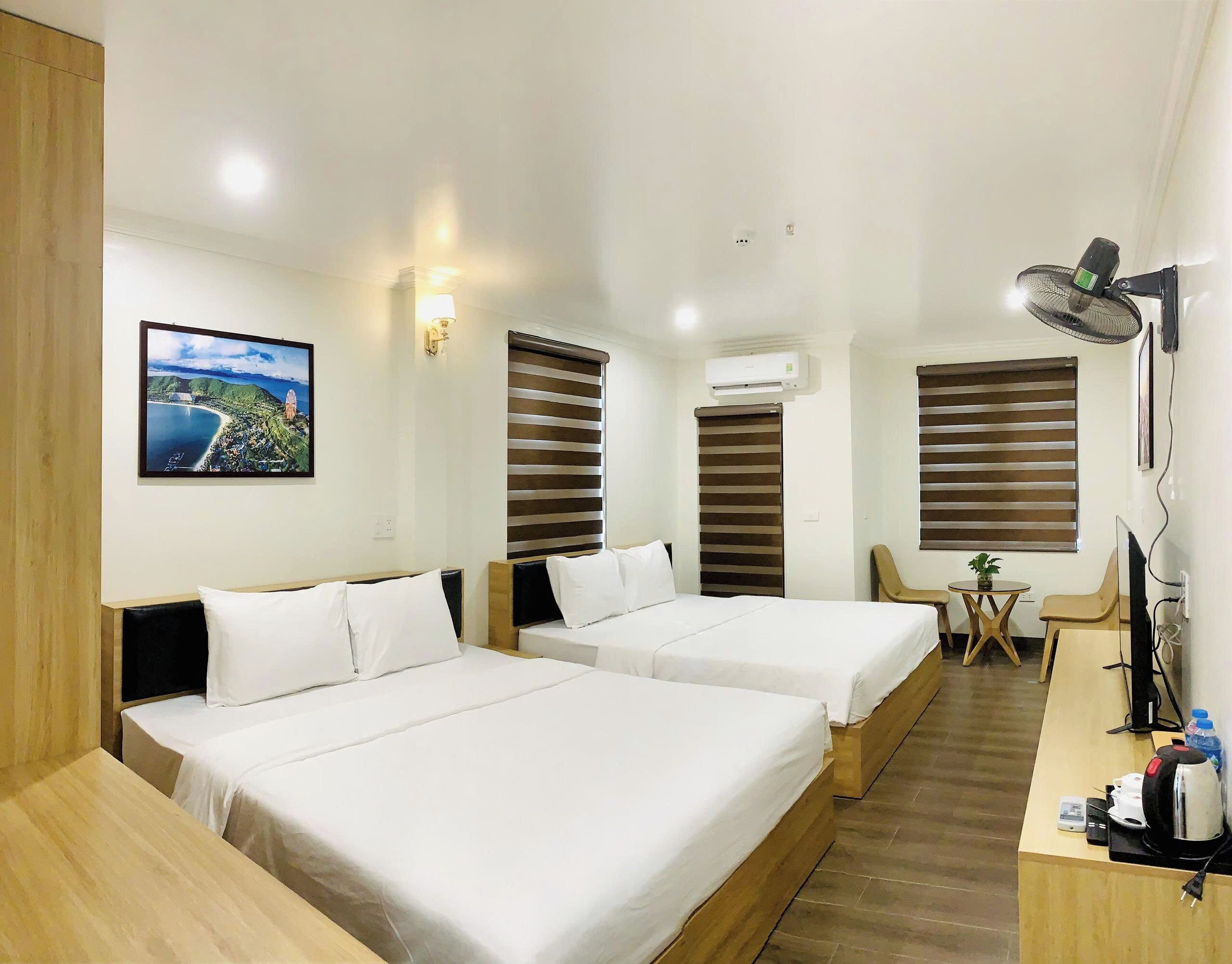 A hotel room with two beds, a desk with a chair, a small table, a lamp, and an air conditioning unit.