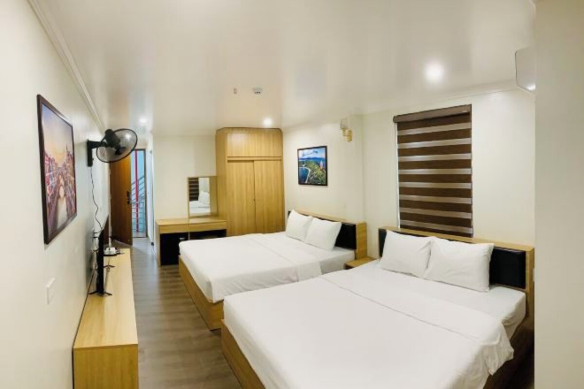 A hotel room with two single beds, a wooden wardrobe, a desk with a chair, and a wall-mounted fan.