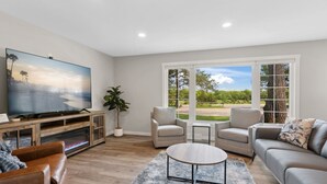 Un salon spacieux doté d’un grand téléviseur à écran plat, de sièges confortables et offrant une vue sur un paysage verdoyant grâce à de grandes fenêtres.