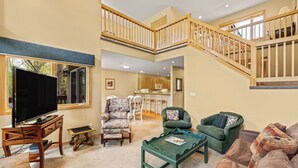 A spacious living area with a high ceiling, a staircase, a flat-screen TV, a dining area, and a kitchen in the background.