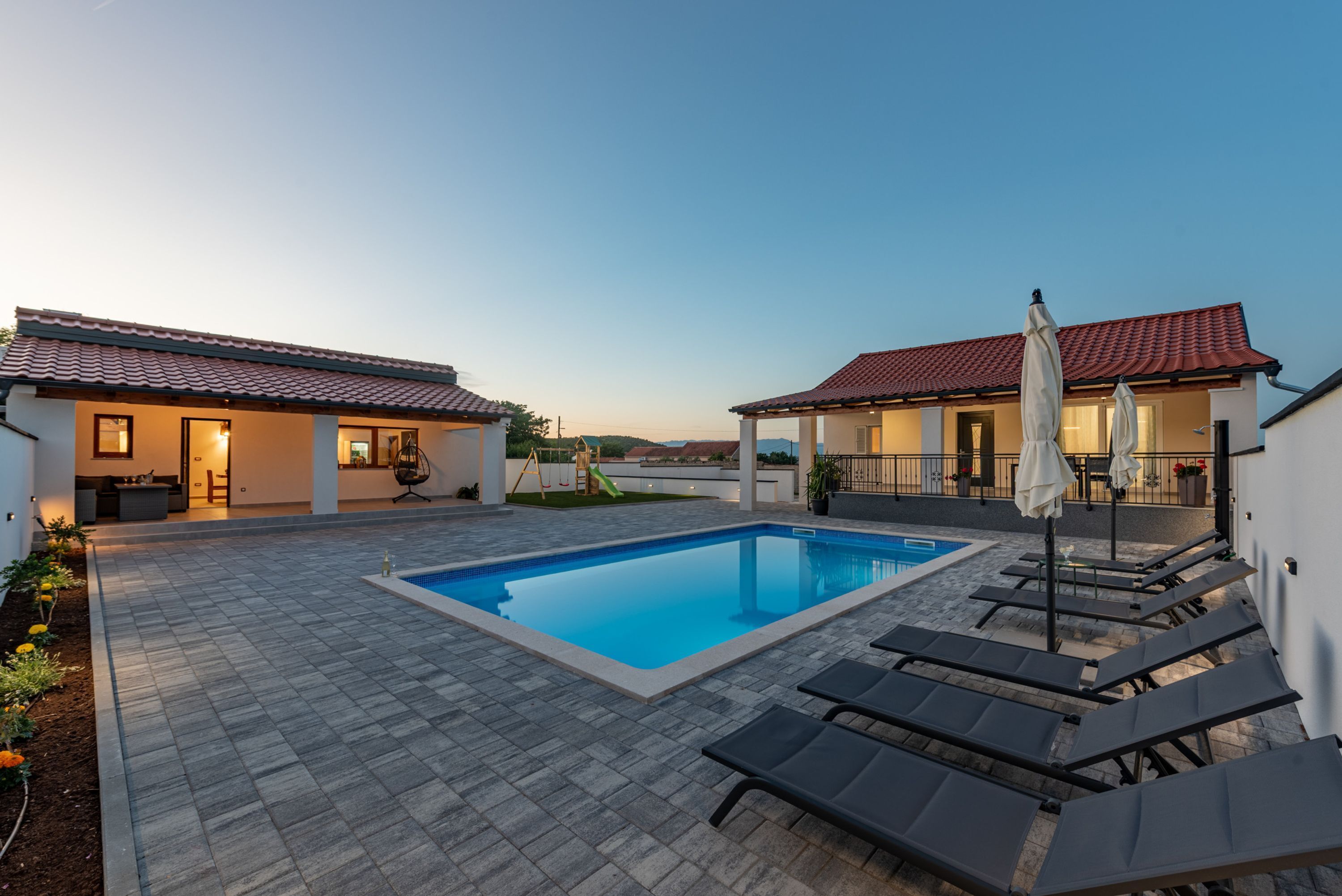 A pool area with lounge chairs and a small slide, surrounded by buildings with red-tiled roofs.