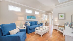 A living room with a blue sofa, a white coffee table, and a wicker chair. There are two lamps on side tables, a painting on the wall, and a window above the sofa. - Updated Cottage Steps to the Beach, Pool and Beach Gear (Holmes Beach)