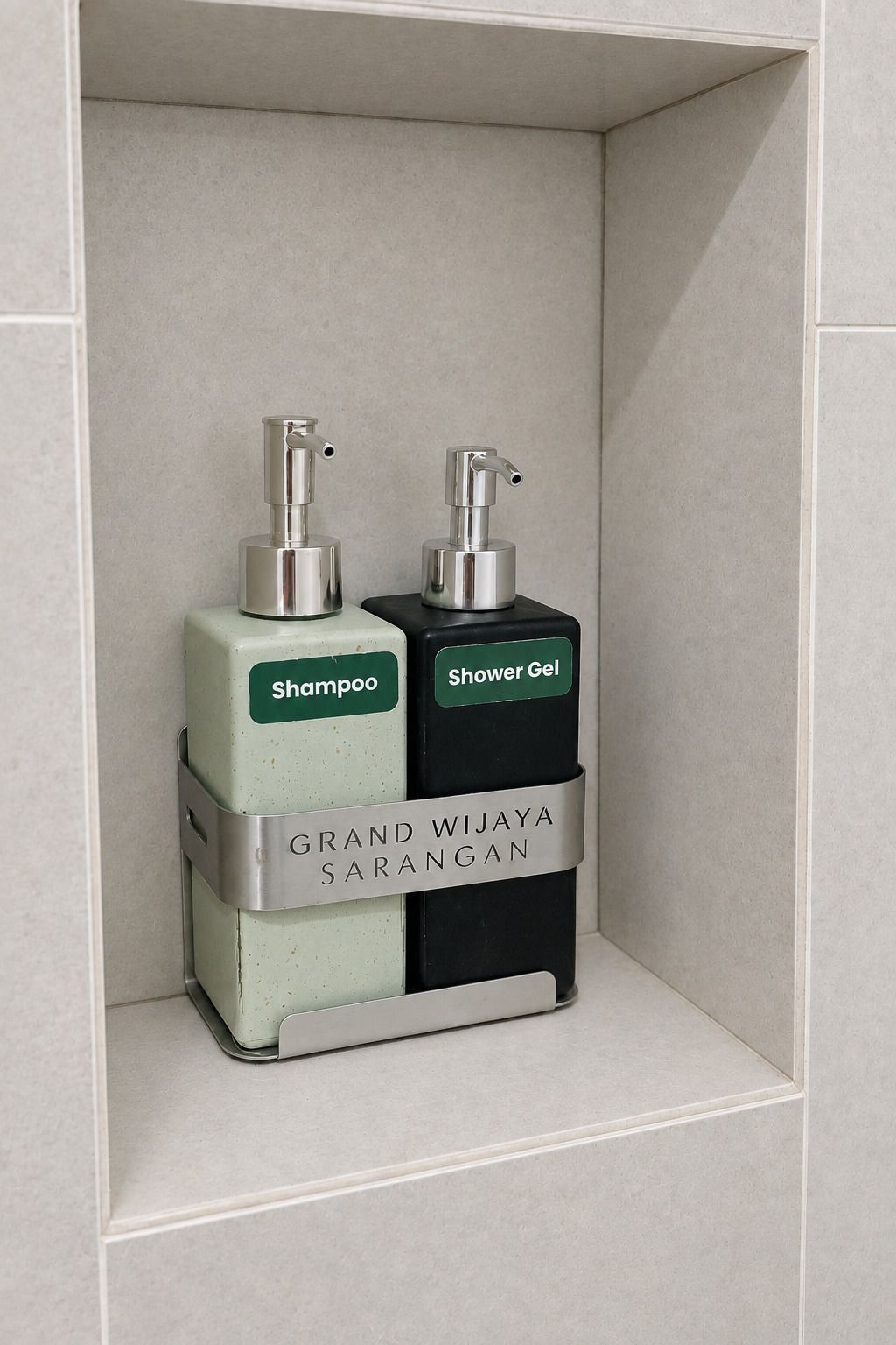Una repisa de baño con dos dispensadores de artículos de tocador con la etiqueta 'Champú' y 'Gel de ducha', y una placa de metal con el texto 'GRAND WIJAYA SARANGAN'.
