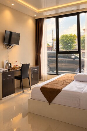 Une chambre d’hôtel comprenant un lit, un bureau, une chaise, une télévision et une grande fenêtre donnant sur la rue.