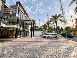 A modern building with a glass facade, a covered entrance, and a paved driveway with parked cars. - Grand Wijaya Hotel dan Villa Sarangan (Plaosan)
