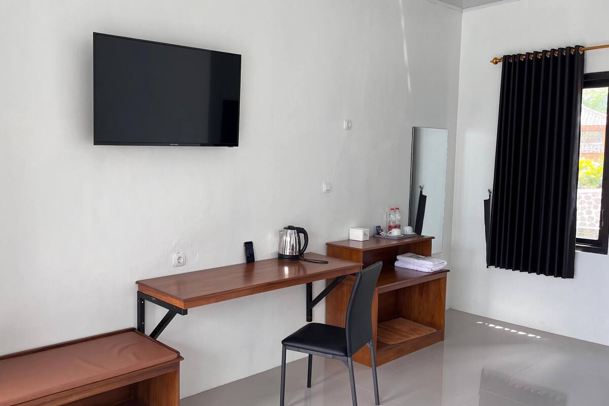 Quarto moderno com televisão de tela plana na parede, escrivaninha de madeira com cadeira, um banco e uma pequena mesa com chaleira e xícaras.