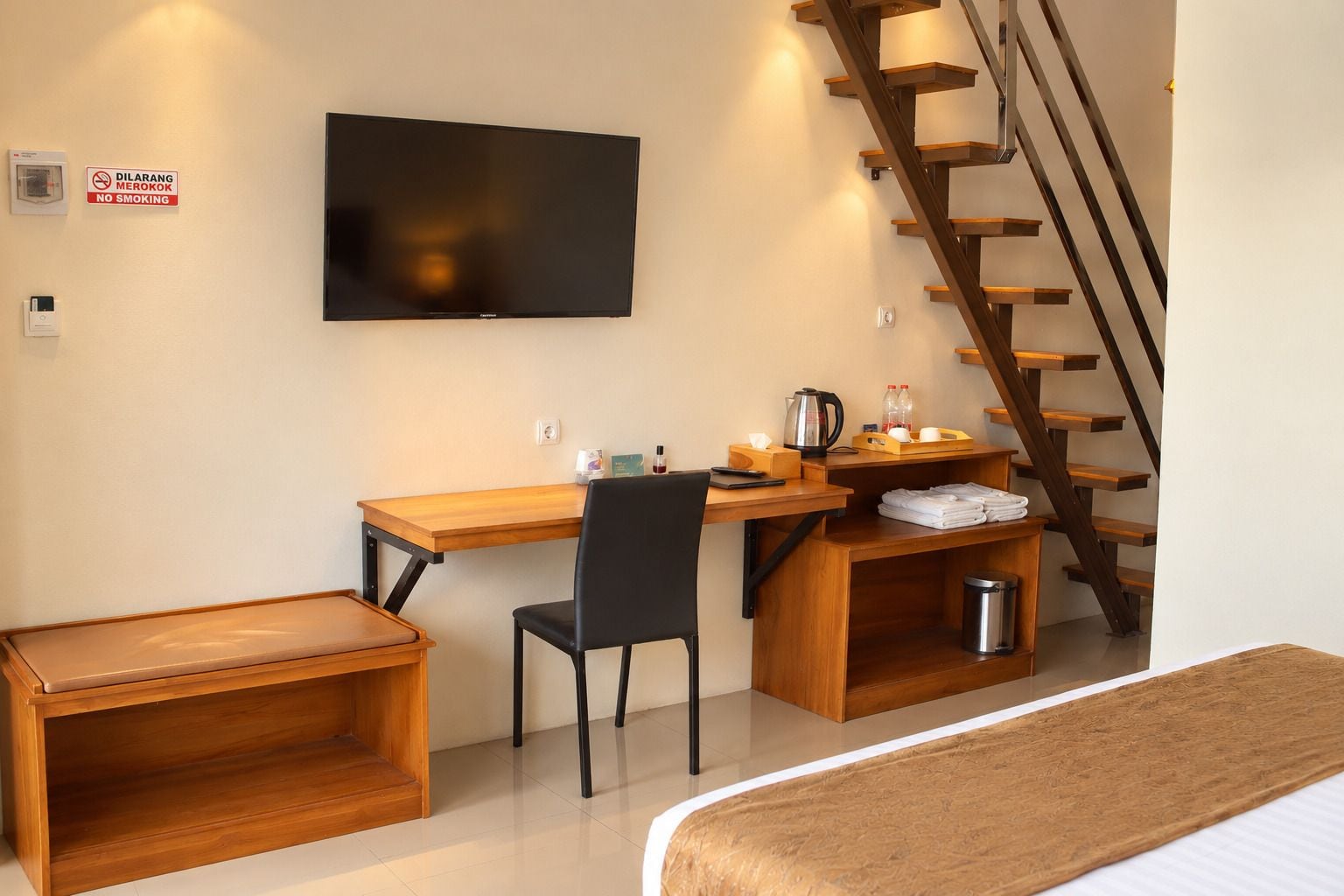 Habitación pequeña con escritorio de madera, una silla, cama, televisor, escalera y un cartel de 'No Fumar'.
