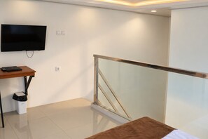 A modern hotel room with a flat-screen TV mounted on the wall, a wooden desk with a laptop, a trash bin, and a glass railing. - Grand Wijaya Hotel dan Villa Sarangan (Plaosan)