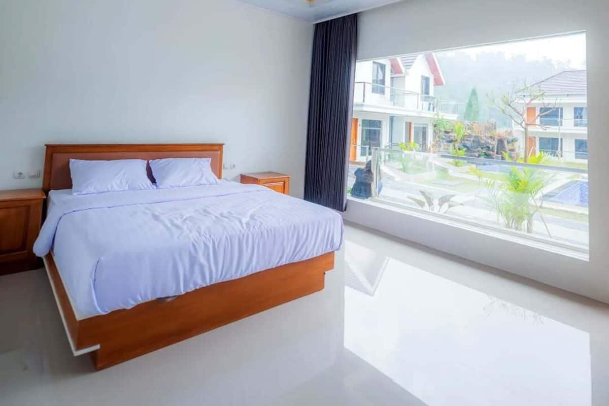 Quarto moderno com cama grande, mesa de cabeceira e janela com vista para edifícios e vegetação.