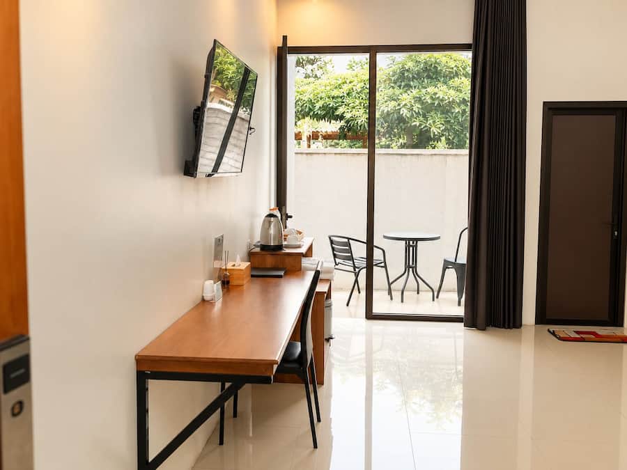 A hotel room with a wooden desk, a flat-screen TV mounted on the wall, a sliding glass door leading to an outdoor seating area with chairs and a table.