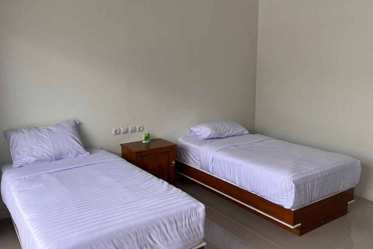 Quarto com duas camas de solteiro, uma mesinha de cabeceira de madeira e uma tomada na parede.