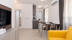 Ein modernes Wohnzimmer mit Flachbildfernseher, einem gelben Sofa, einem Essbereich mit Tisch und Stühlen und einer Küche im Hintergrund.
