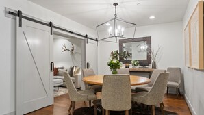 A dining area with a round wooden table, six grey upholstered chairs, a chandelier, and a cowhide rug.