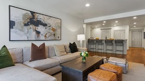 A modern living room with a sofa, coffee table, and ottomans. There is a kitchen area with bar stools and a large abstract painting on the wall.