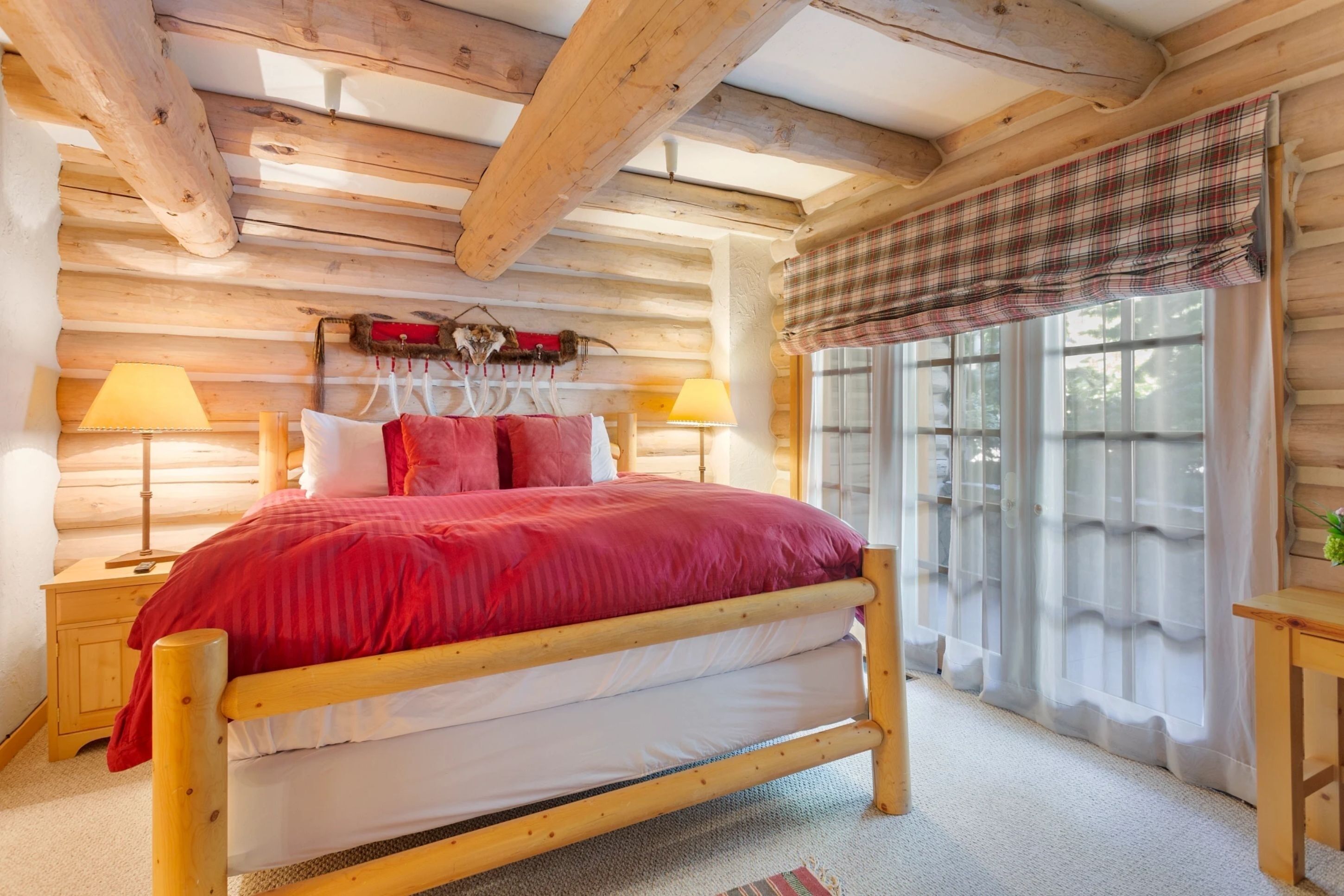 A cozy log cabin bedroom with a wooden bed, two bedside lamps, and a plaid window valance.