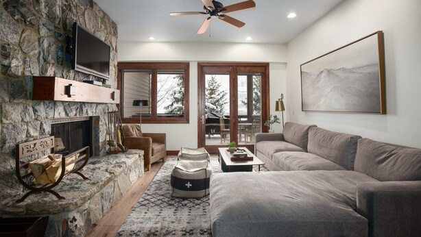 A cozy living room with a stone fireplace, a flat-screen TV, a ceiling fan, and a large window with a view of snow-covered trees. - Trail's End Lodge at Deer Valley Resort - Two Bedroom Residence with Spa #403 2 Condo (Park City)