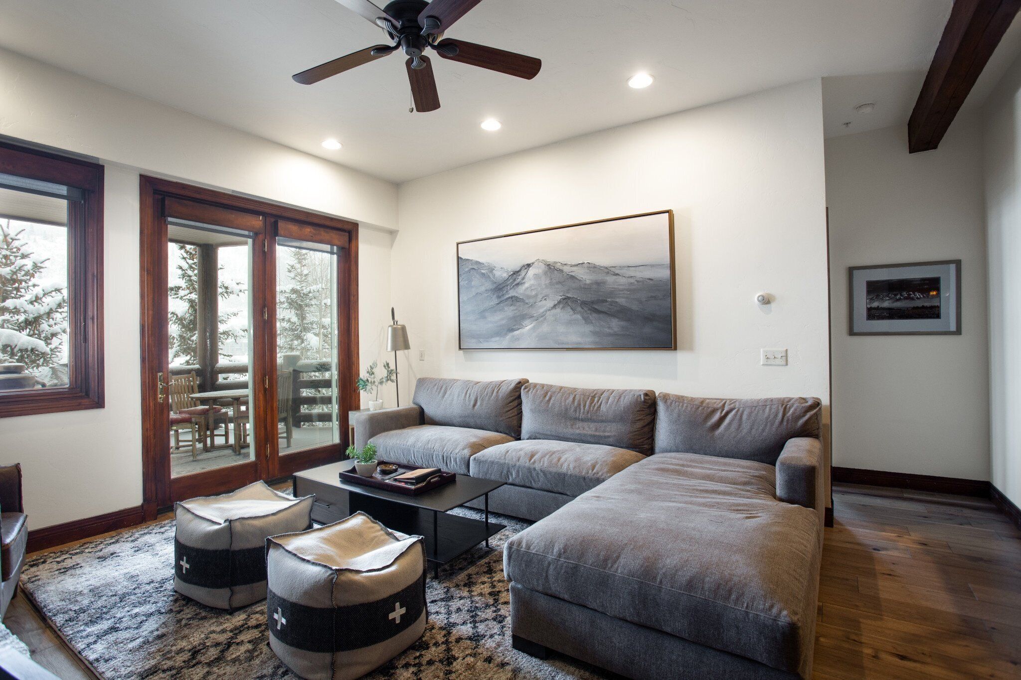 A modern living room with a sectional sofa, a coffee table, and ottomans. There is a ceiling fan, a large window, and a painting on the wall.