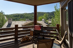 Una terraza de madera con una parrilla, sillas y vistas a una ladera boscosa.