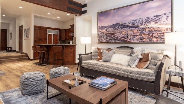 A modern living room with a sofa, ottomans, a coffee table, and a large painting of a snowy mountain town. - Trail's End Lodge at Deer Valley Resort - Two Bedroom Residence with Spa #402 2 Condo (Park City)