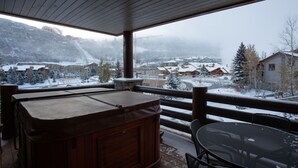 Terraza de madera con un jacuzzi y una mesa, con vistas a un pueblo nevado con casas y montañas.
