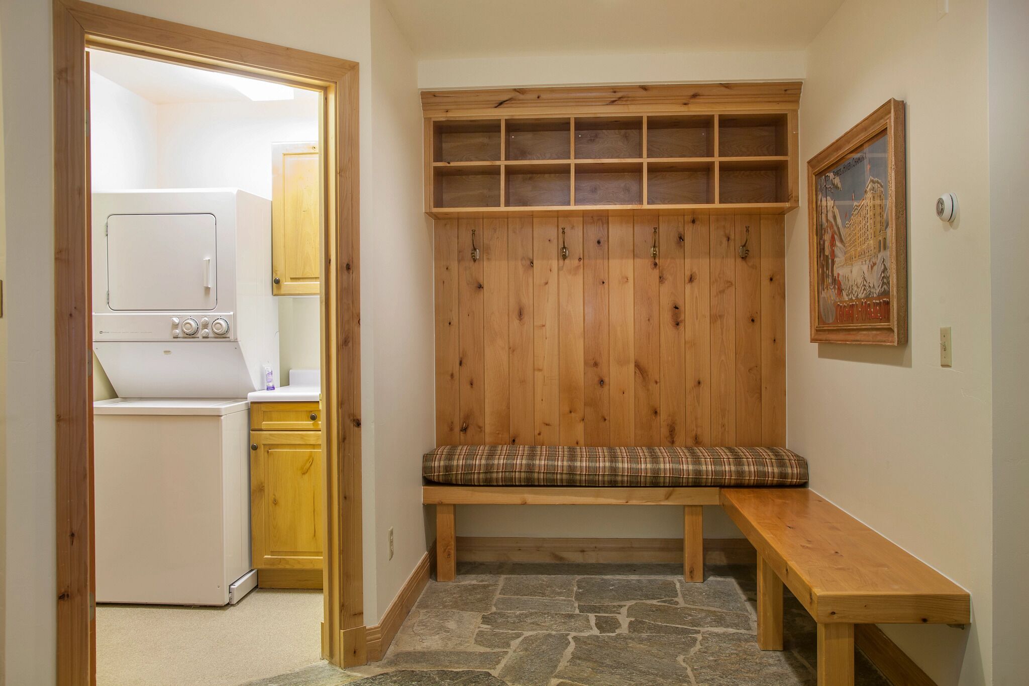 A laundry room with a washing machine, a bench with a plaid cushion, and a wooden shelving unit.