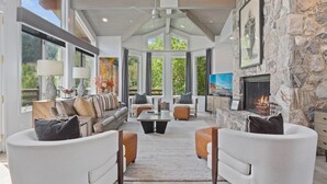 A spacious living room with a stone fireplace, large windows, and a high ceiling.