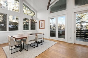 Une salle à manger avec une table et des chaises en bois, de grandes fenêtres et un lustre.