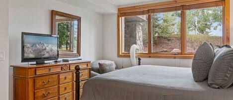 Un dormitorio con cama, cómoda, televisor y ventana con vista a árboles.
