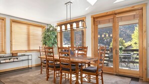 A dining area with a wooden table and chairs, a hanging light fixture, and large windows with wooden blinds.