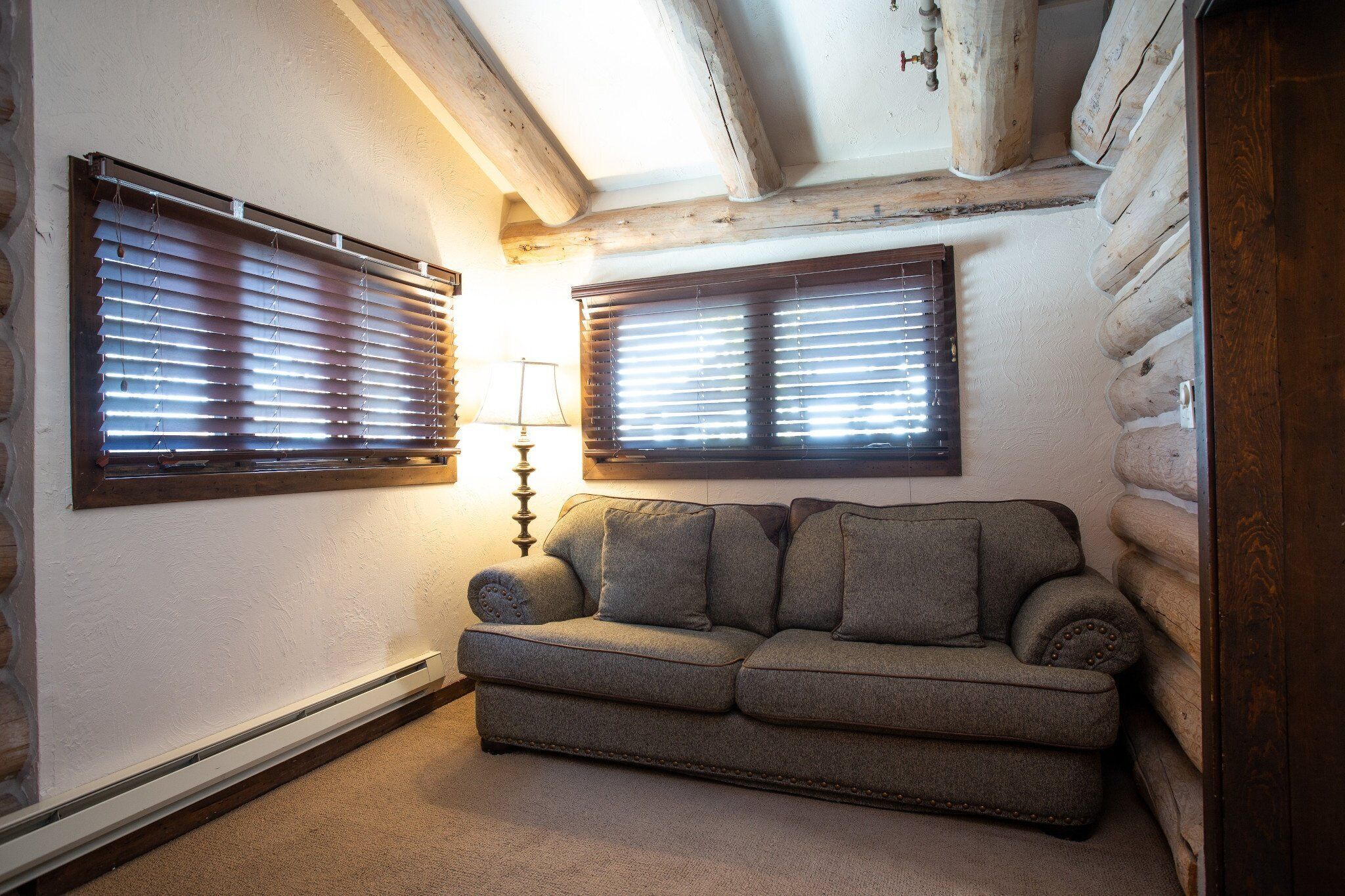 Una habitación acogedora con sofá, dos ventanas con persianas, una lámpara y techo de madera.
