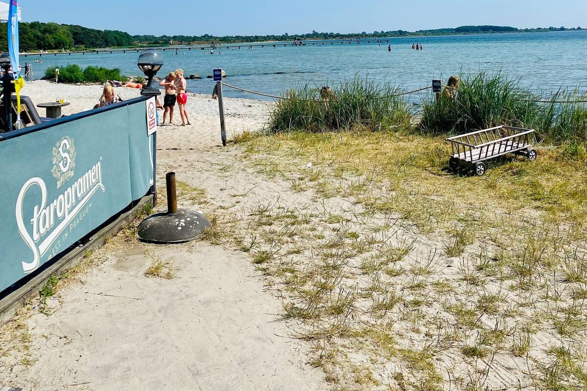 Ein Strand mit einem Schild für Staropramen, eine sandige Fläche mit Gras und einem Gewässer im Hintergrund.