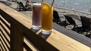 Two drinks on a wooden deck overlooking a beach with lounge chairs and a view of the water.
